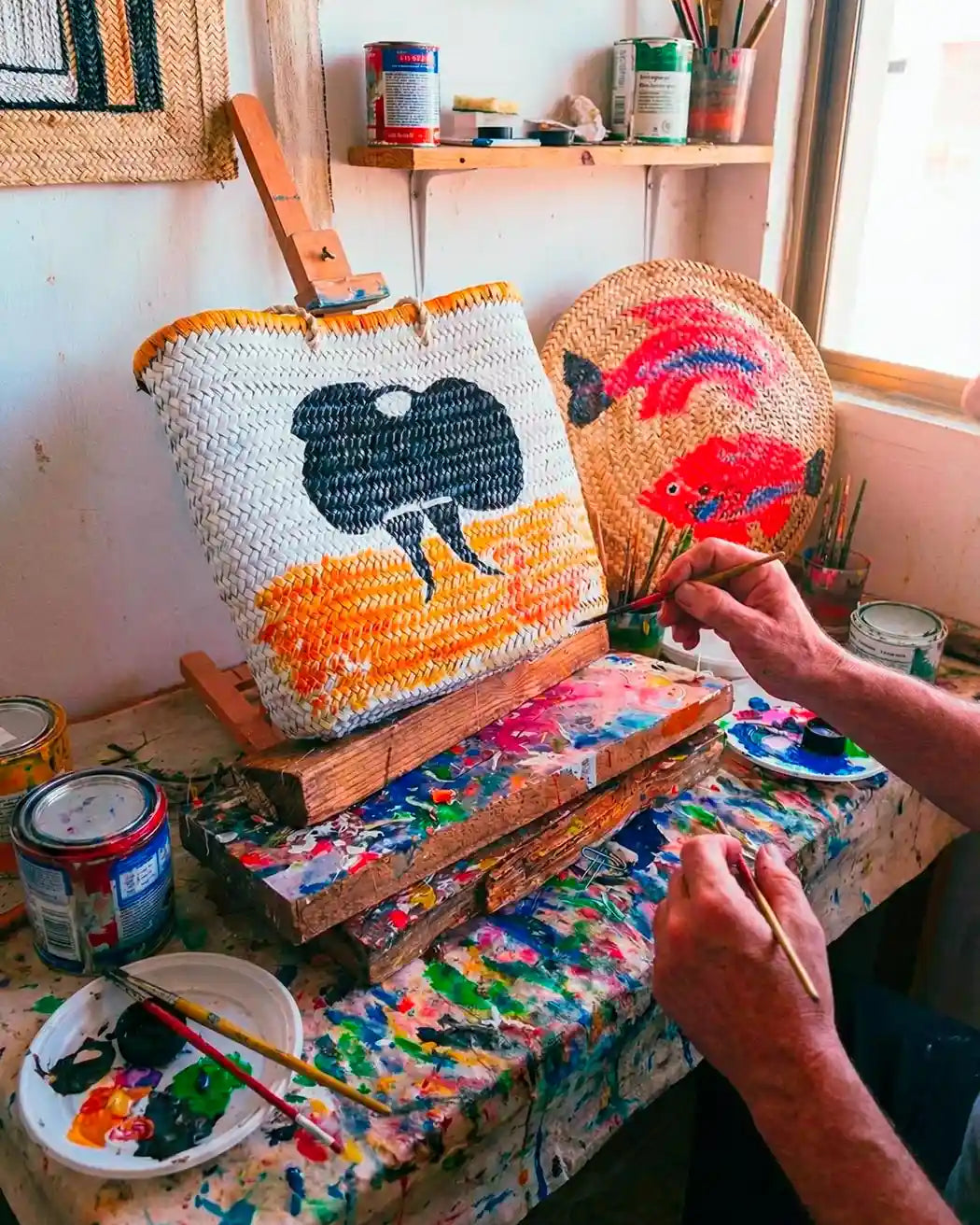 Scorcio luminoso e colorato dell'atelier dell'artista: un banco da lavoro vissuto, colmo di pennelli, colori e attrezzi del mestiere, fa da cornice a una parete dove sono esposte borse di paglia uniche, decorate con vivaci motivi dipinti a mano.