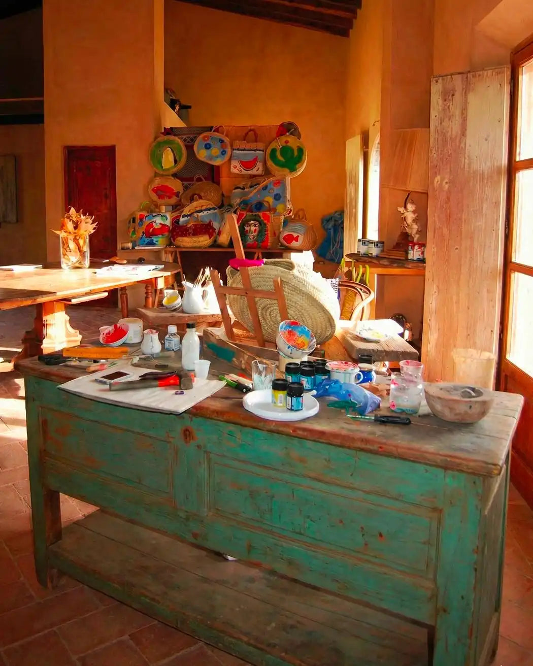 Foto verticale di un laboratorio artistico rustico e inondato di sole. In primo piano, un grande banco da lavoro in legno verde decapato è coperto di barattoli di vernice, pennelli, ciotole e attrezzi. Al centro, un cavalletto regge una borsa di paglia in lavorazione. Sullo sfondo, una parete decorata con numerose borse di paglia dipinte e colorate, travi in legno a vista sul soffitto e una grande finestra sulla destra che lascia entrare luce calda.