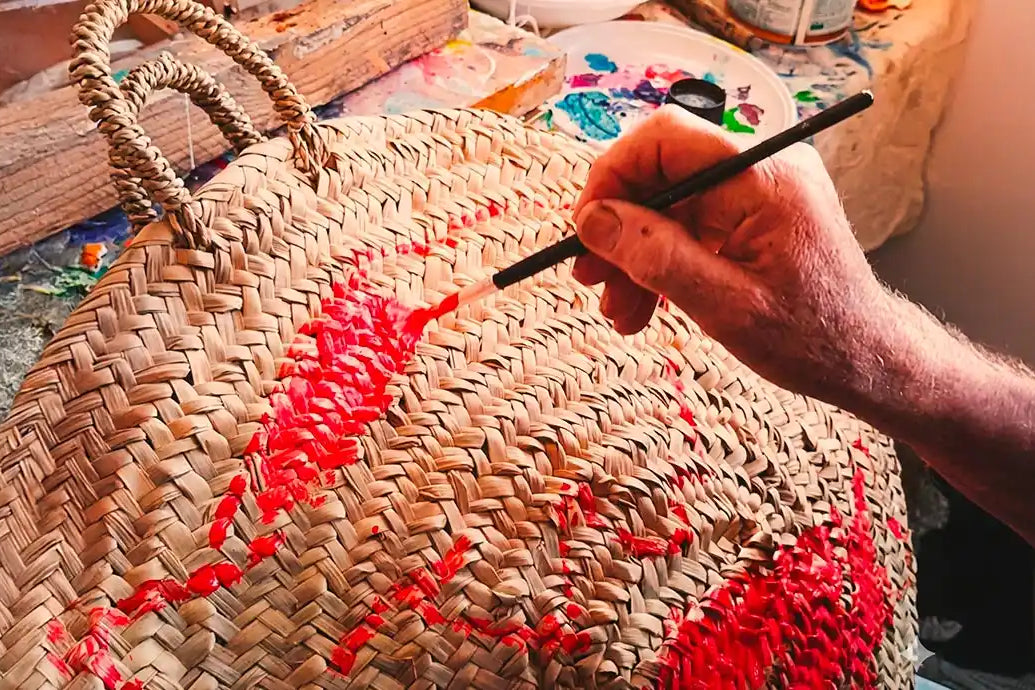 Primo piano delle mani di un artigiano che dipingono con cura un motivo rosso vibrante su una borsa di paglia intrecciata, utilizzando un pennello sottile all'interno del suo laboratorio creativo.