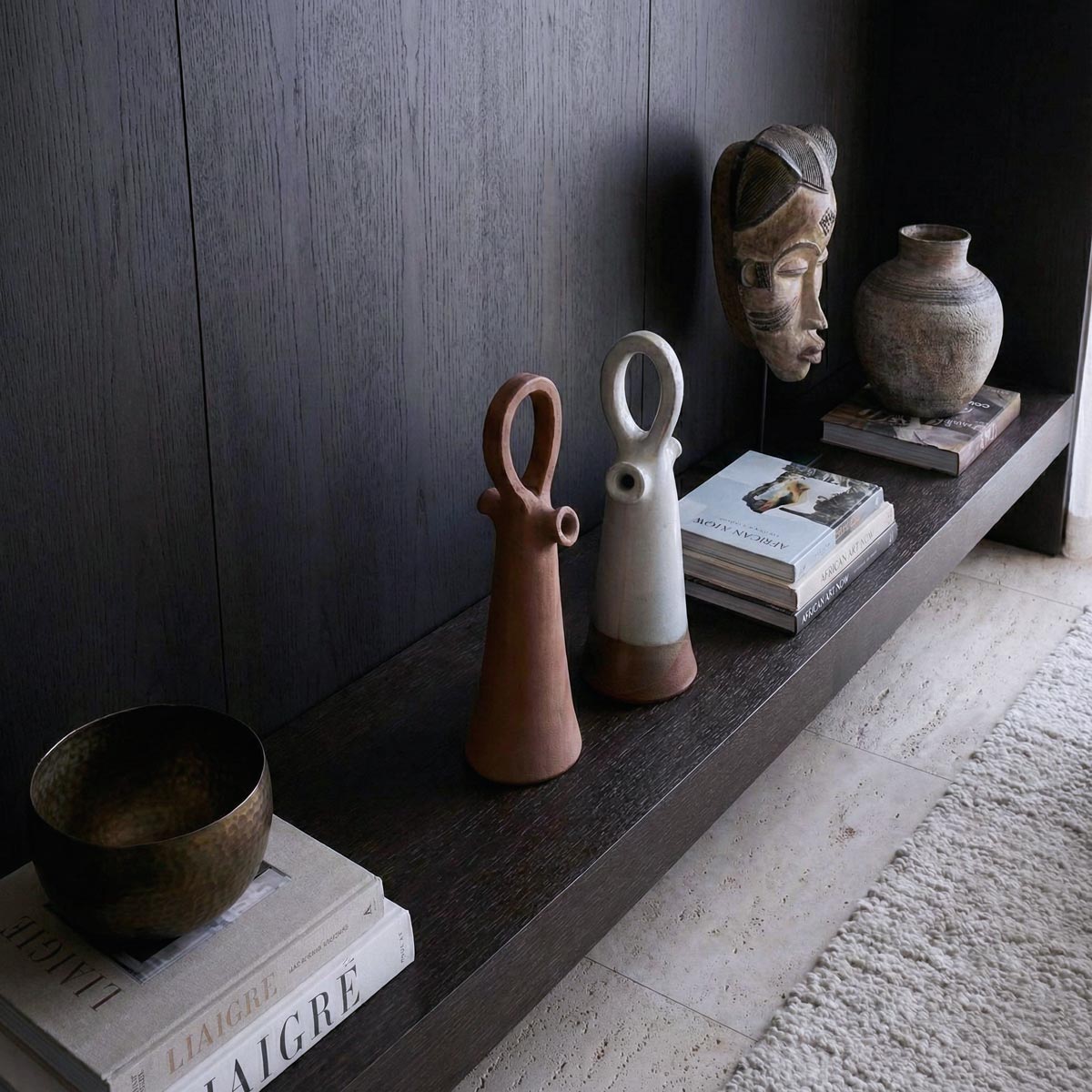 Sofisticata ambientazione di interior design in stile minimale e materico. Su una console in legno scuro spiccano due vasi scultura in ceramica (terracotta e smaltato), circondati da una selezione curata di oggetti d'arte, libri di design e manufatti etnici. L'illuminazione naturale laterale esalta le texture dei materiali naturali.