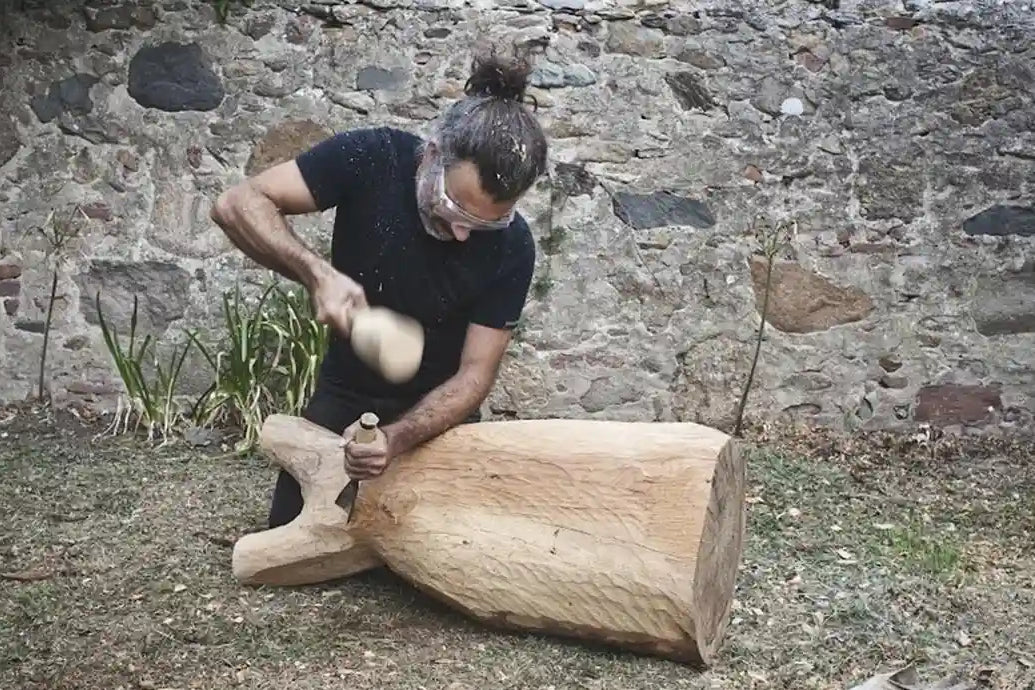 Estebanland seduto nel suo laboratorio all'aperto, circondato da sculture in legno in lavorazione.