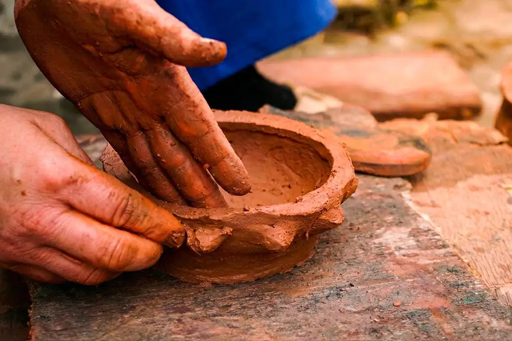 Dettaglio delle mani di un artigiana tunisina che modellano argilla rossa grezza su un tavolo da lavoro rustico, rappresentando la tradizione della ceramica fatta a mano malles per vicode