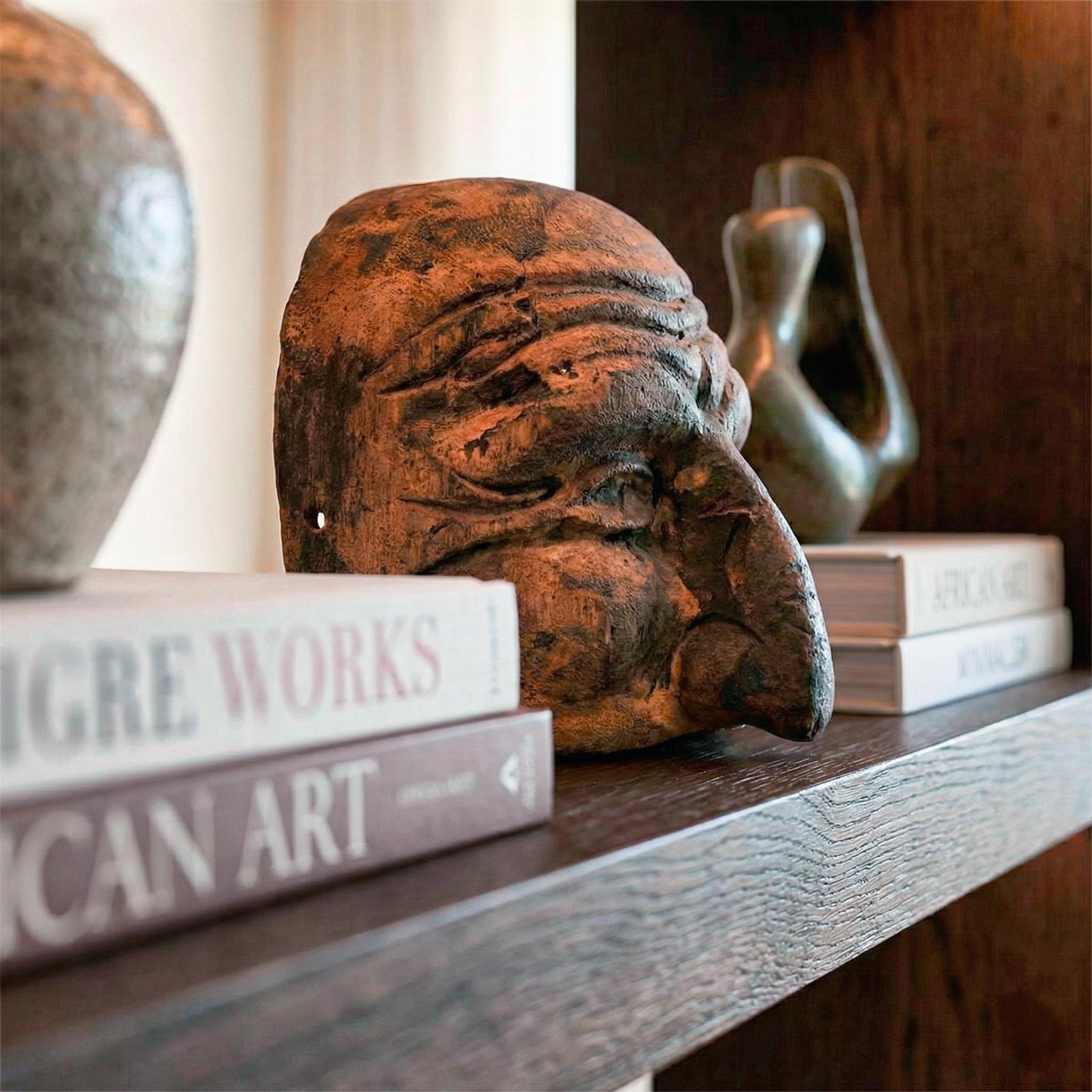 Primo piano con taglio prospettico laterale dell'antica maschera di Pulcinella in legno. L'inquadratura ravvicinata evidenzia la texture invecchiata e le venature del legno scuro della scultura, posizionata su una mensola accanto ai dorsi dei libri 'Liaigre Works' e 'African Art'. Lo sfondo è sfocato.
