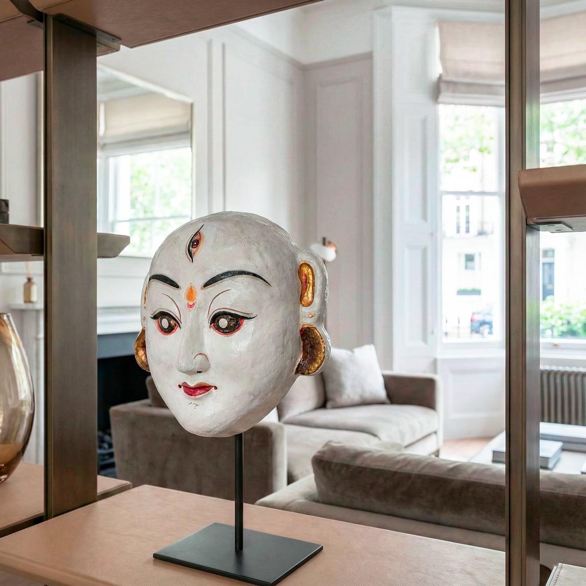 Primo piano artistico della maschera 'Ballo di Durga'. La ripresa ravvicinata evidenzia la texture artigianale della cartapesta bianca e i dettagli dipinti a mano del volto sacro. La scultura, alta 18 cm, si staglia nitida sul supporto in metallo nero, mentre lo sfondo sfocato lascia intuire un ambiente d'interni lussuoso e moderno.