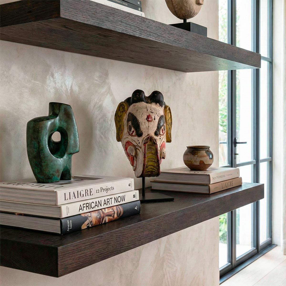 Zoom ravvicinato con prospettiva dal basso di una maschera tribale in legno intagliato e dipinto a mano, che mostra la texture invecchiata della vernice bianca, rossa e nera. La maschera è posizionata sopra i libri d'arte 'LIAIGRE 12 Projects' e 'AFRICAN ART NOW', i cui dorsi sono visibili in primo piano. Lo sfondo con la mensola e la finestra è sfocato.