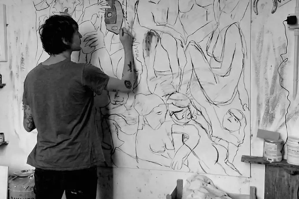 L'artista Massimiliano Precisi di spalle nel suo studio che dipinge su una grande tela piena di figure stilizzate e volti, circondato da altri quadri appesi e appoggiati.