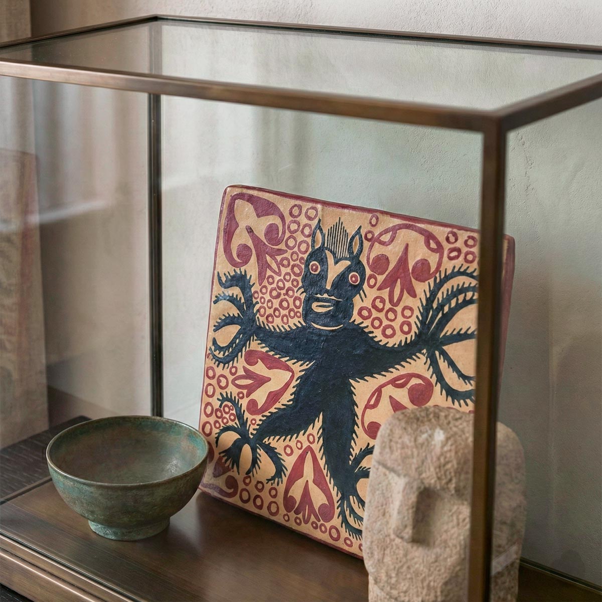 Primo piano ravvicinato all'interno di una teca espositiva in vetro e ottone. Il focus è sulla texture di una mattonella quadrata in ceramica dipinta a mano, che raffigura un diavolo nero stilizzato tra motivi rossi. In primo piano, parzialmente visibili, una ciotola in bronzo patinato e la testa di una scultura in pietra.