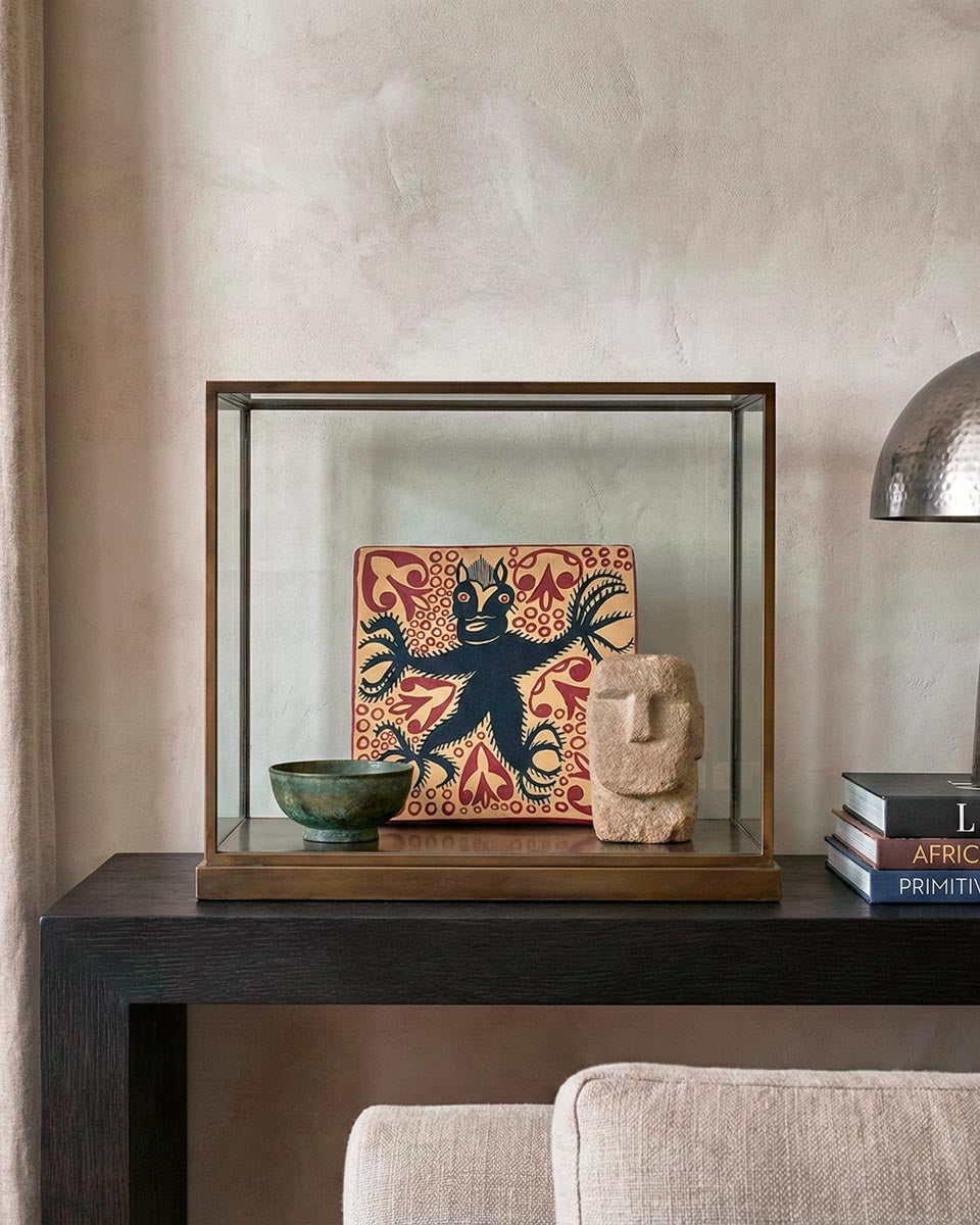 Lussuosa composizione di interior design in stile Liaigre. Su una console scura, una teca in vetro con telaio in ottone espone una mattonella in ceramica decorata con un diavolo nero su fondo rosso, affiancata da una ciotola in bronzo e una scultura Moai in pietra. A lato, una pila di libri di design (tra cui 'Liaigre' e 'African Art') e una lampada in metallo martellato.