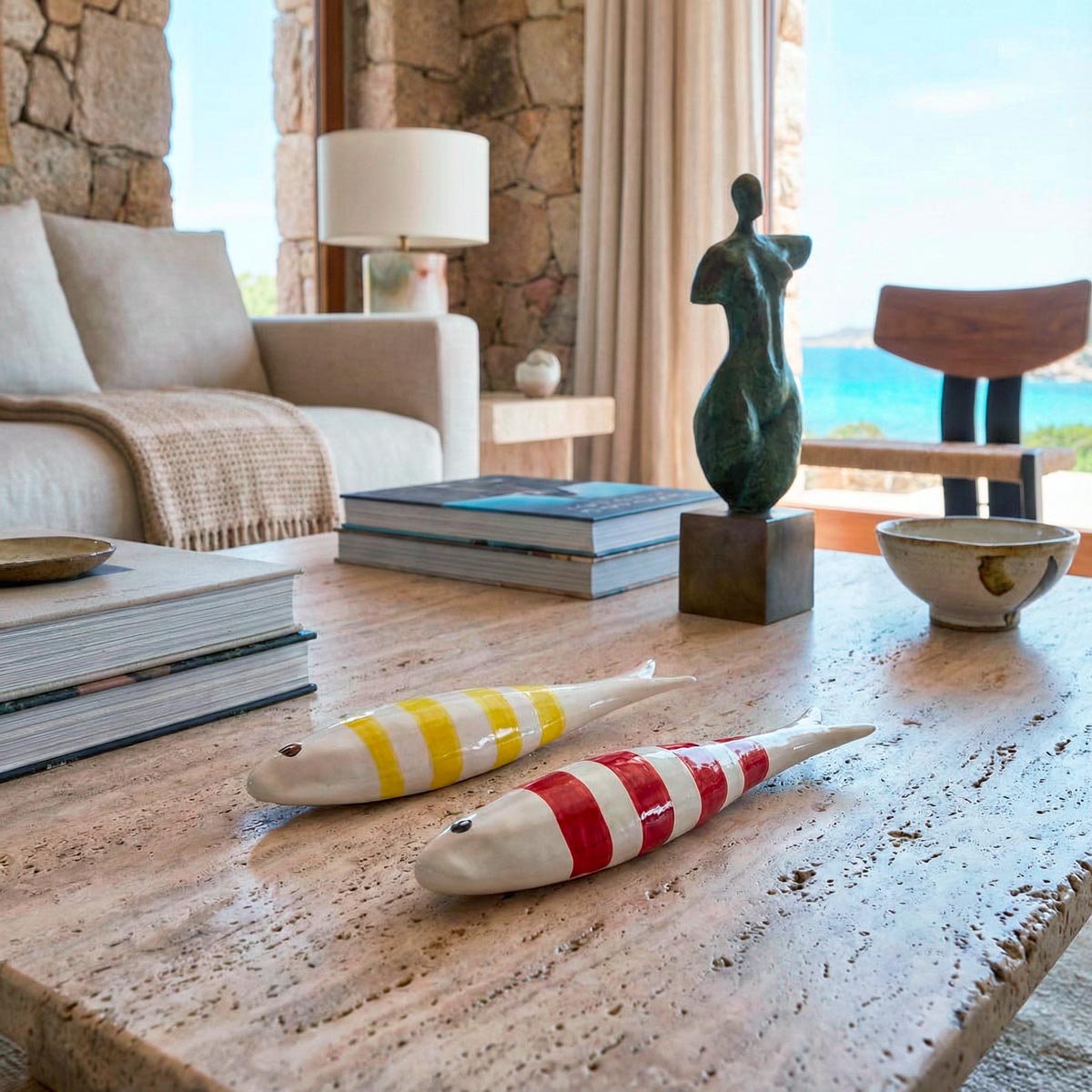Primo piano con angolazione laterale di un tavolino da salotto in pietra grezza. Il focus è su due sculture in ceramica a forma di pesce, lunghe circa 8 cm, decorate con righe bianche e colorate (giallo e rosso). La luce naturale ne esalta la finitura lucida, mentre sullo sfondo sfocato si intravedono arredi moderni e il mare.