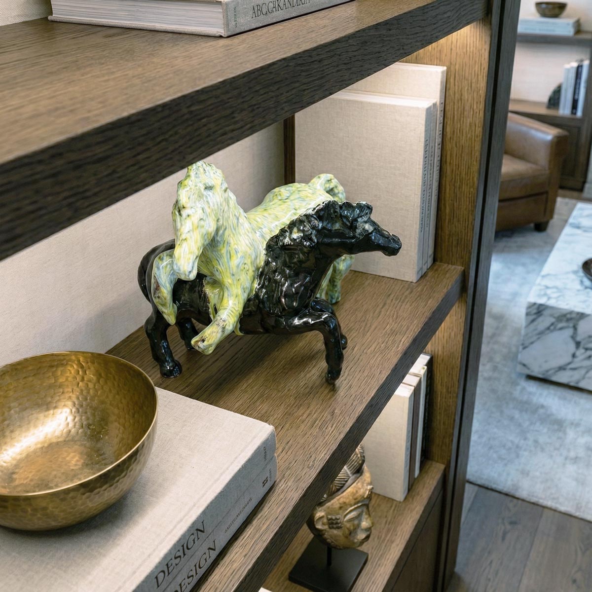 Dettaglio ravvicinato di una scultura decorativa in ceramica lucida con due cavalli in movimento. L'inquadratura dall'alto mette in risalto la finitura marmorizzata gialla e verde del primo cavallo e quella nera del secondo. L'oggetto è posizionato su una mensola in rovere illuminata, accanto a pile di libri d'arte 'Design'.