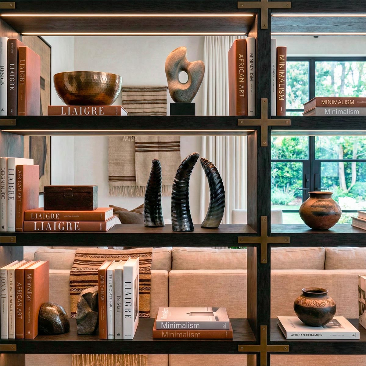 Libreria di alta ebanisteria in legno scuro e dettagli in bronzo, situata in un living lussuoso in stile Liaigre. Sui ripiani illuminati a LED è esposta una collezione curata di oggetti, con al centro tre sculture a forma di corno in osso nero marocchino, affiancate da libri di design come 'Liaigre' e 'Minimalism', una ciotola in bronzo martellato e sculture in pietra. Sullo sfondo, un salotto con divano in lino e vetrate sul giardino.