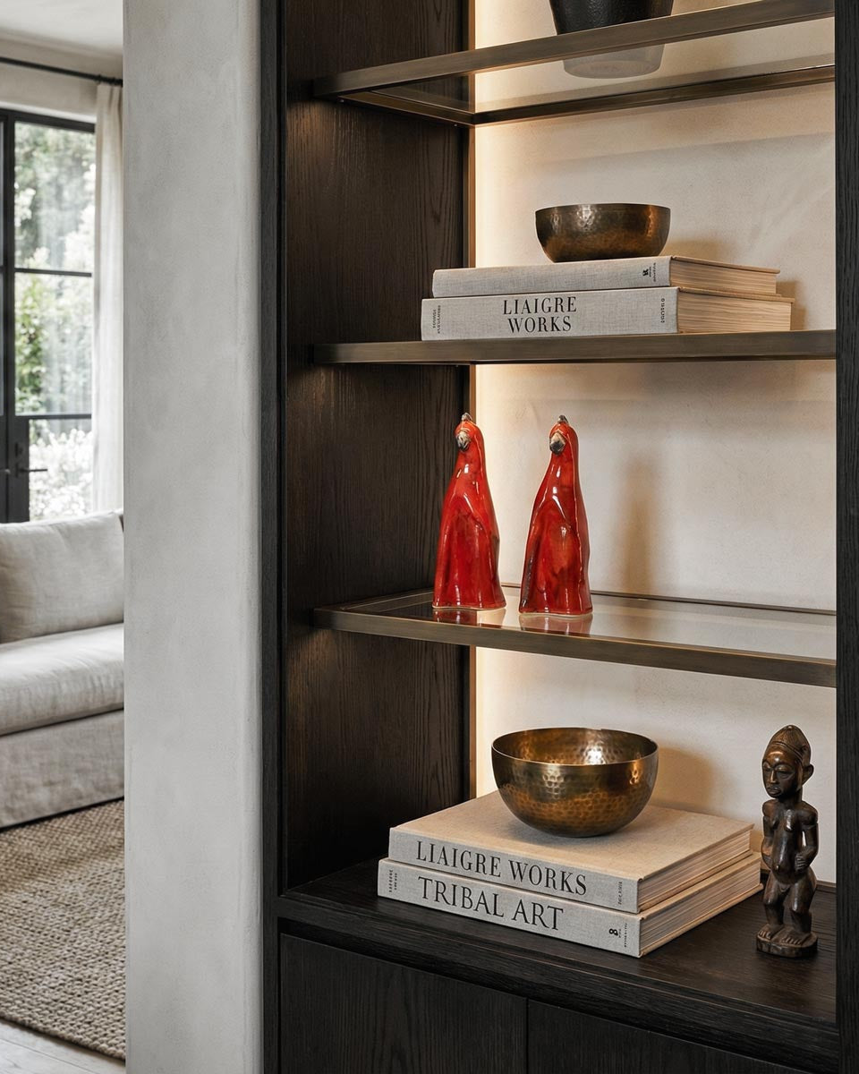 Coppia di sculture artigianali in ceramica rossa a forma di pappagallo, esposte come accento di colore su una mensola in vetro e bronzo illuminata. La libreria a muro in legno scuro stile Liaigre contiene anche libri d'arte ('Liaigre Works', 'Tribal Art'), ciotole in bronzo e una statuetta etnica, in un salotto luminoso e sofisticato