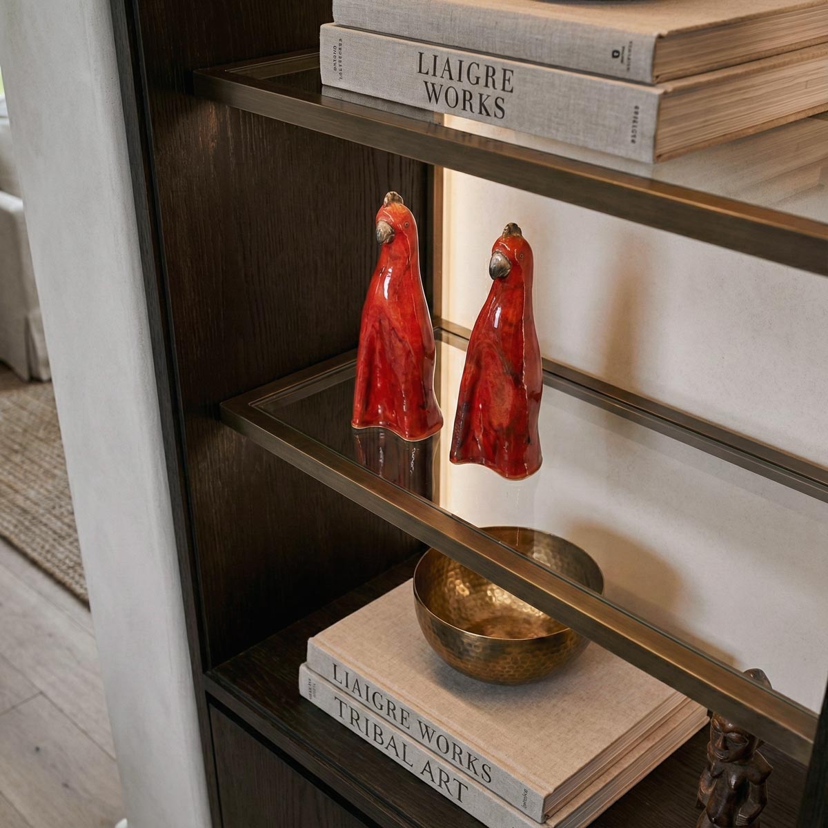 Dettaglio ravvicinato dall'alto delle due sculture di pappagalli in ceramica. L'inquadratura mette in risalto la finitura rossa lucida e la forma stilizzata degli oggetti, posizionati su un ripiano in vetro con cornice in bronzo. I riflessi della luce LED integrata sono visibili sulla superficie vetrata.