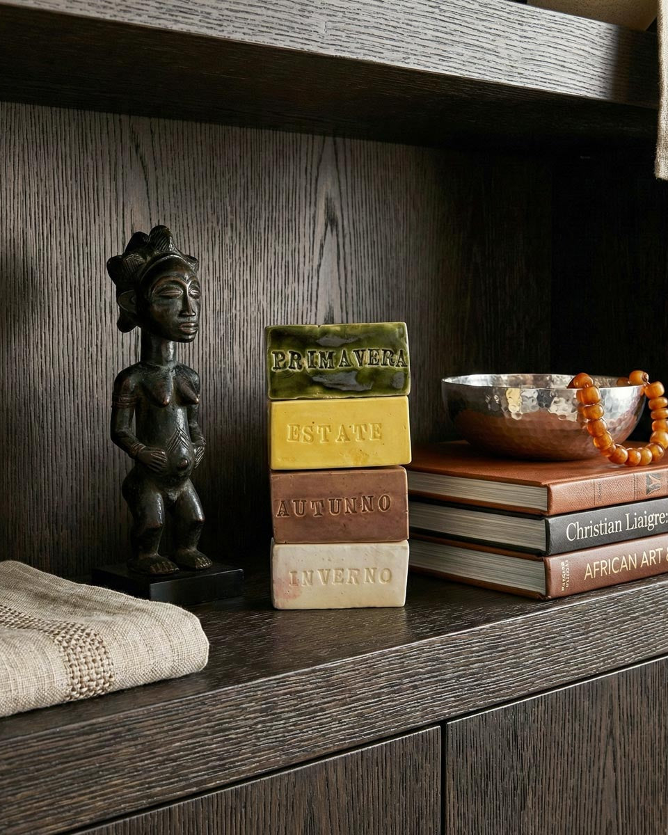 Primo piano di una composizione di design con quattro mattoncini di ceramica smaltata impilati, ognuno con il nome di una stagione in rilievo: PRIMAVERA (verde), ESTATE (giallo), AUTUNNO (marrone) e INVERNO (bianco). A sinistra, una scultura africana in legno scuro raffigurante una figura femminile. Sullo sfondo, una ciotola in metallo martellato con una collana di perle ambrate e dei libri, il tutto su un ripiano di boiserie in rovere scuro in un ambiente di lusso in stile Liaigre.