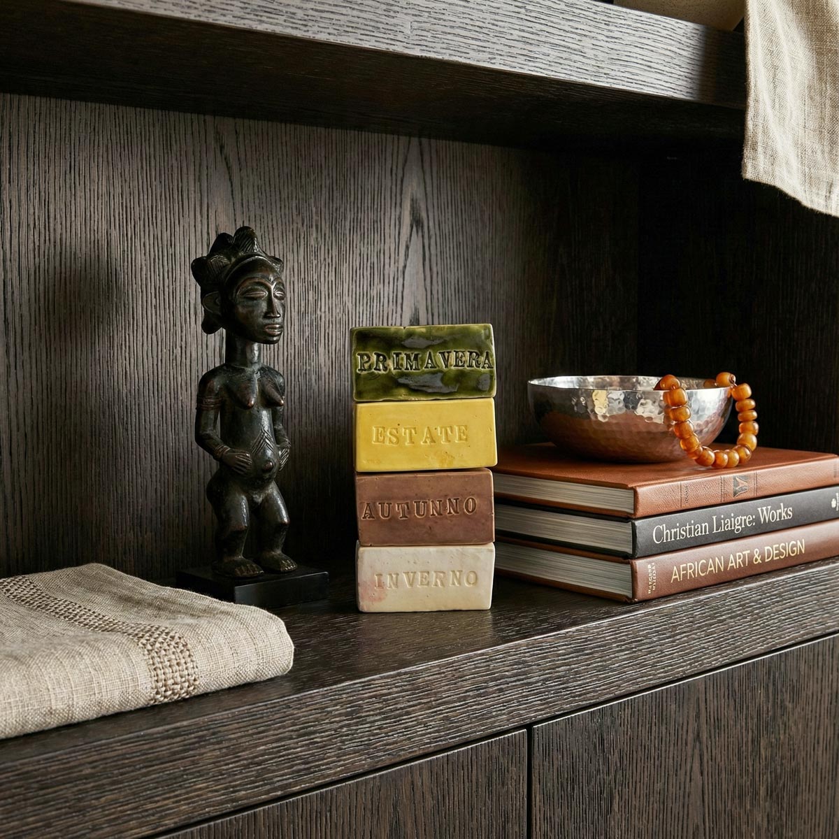 Primo piano di una composizione di design con quattro mattoncini di ceramica smaltata impilati, ognuno con il nome di una stagione in rilievo: PRIMAVERA (verde), ESTATE (giallo), AUTUNNO (marrone) e INVERNO (bianco). A sinistra, una scultura africana in legno scuro raffigurante una figura femminile. Sullo sfondo, una ciotola in metallo martellato con una collana di perle ambrate e dei libri, il tutto su un ripiano di boiserie in rovere scuro in un ambiente di lusso in stile Liaigre.