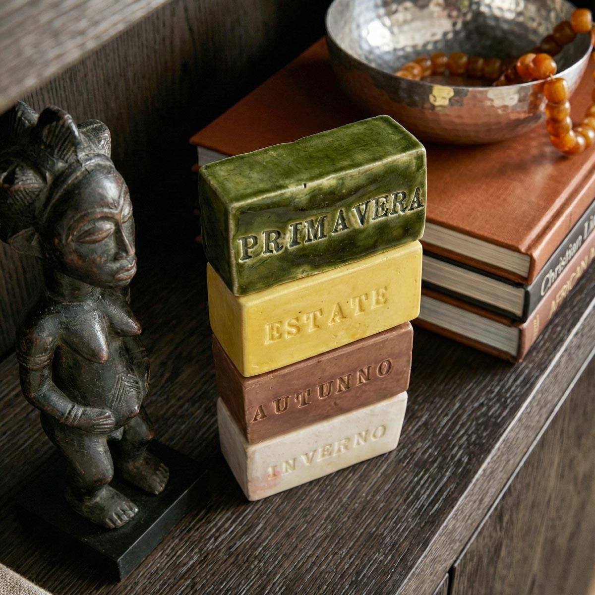 Immagine da rivista di architettura che mostra quattro ceramiche smaltate con i nomi delle stagioni impilate su una mensola di legno scuro, accanto a una statuetta etnica e una ciotola di metallo con perline, in un ambiente di design sofisticato.