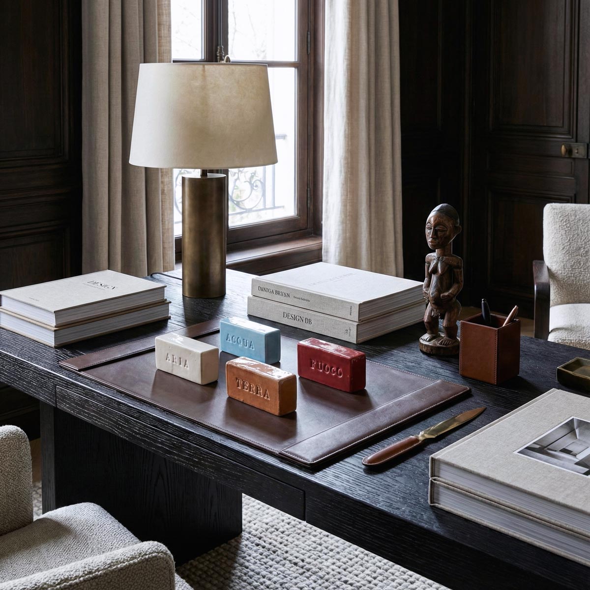 Set di quattro mattoncini in ceramica smaltata con i nomi degli elementi (ARIA, ACQUA, TERRA, FUOCO), disposti su un sottomano in pelle marrone su una scrivania direzionale in legno scuro. L'ambiente lussuoso include una lampada in ottone, libri di design e una scultura africana in legno vicino a una finestra.