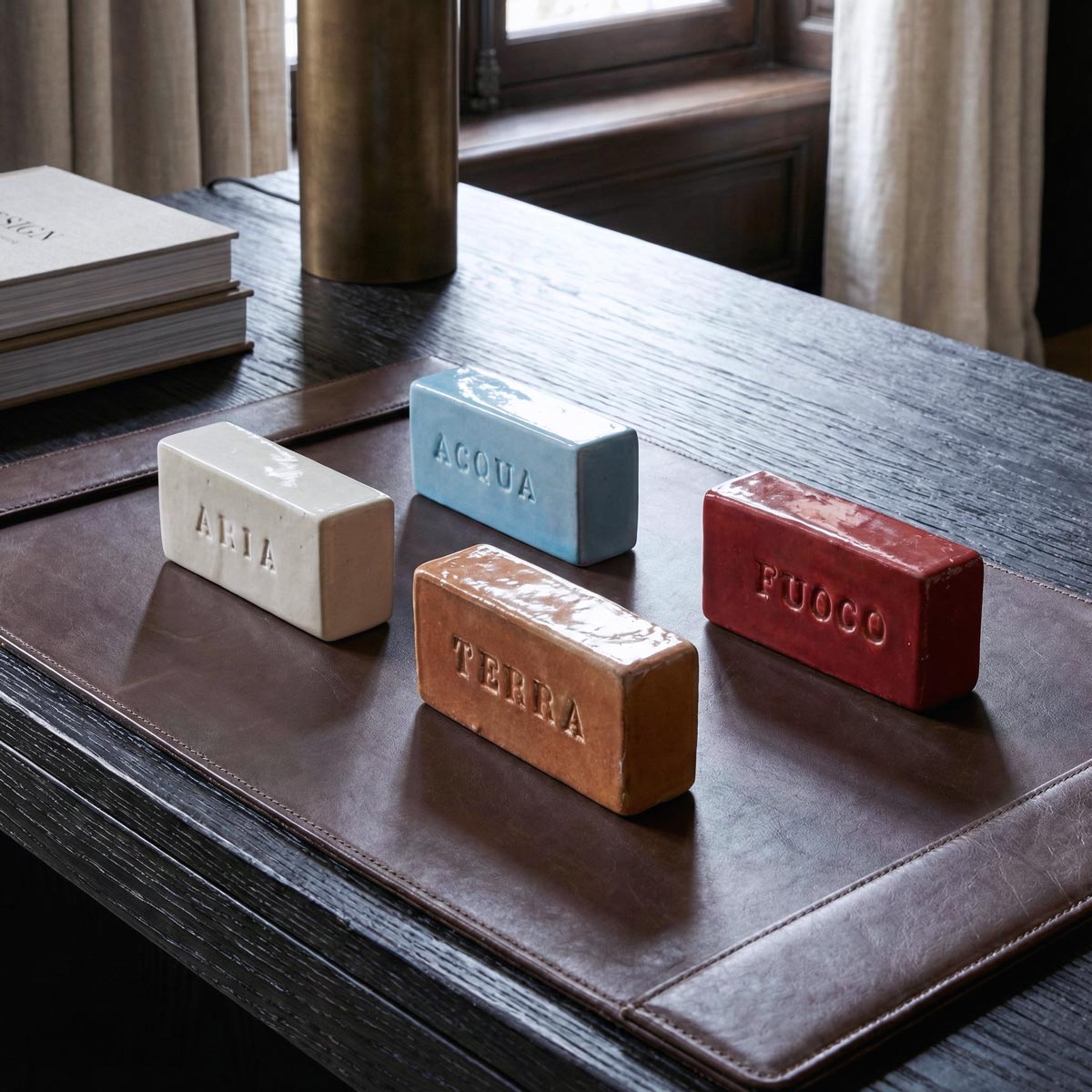 Dettaglio ravvicinato dall'alto dei quattro mattoncini 'Elementi' in ceramica lucida. L'inquadratura evidenzia la texture dello smalto e le scritte incise sui blocchi (bianco, azzurro, terracotta, rosso), appoggiati su un sottomano in pelle con cuciture a vista e la base di una lampada in ottone sfocata sullo sfondo.