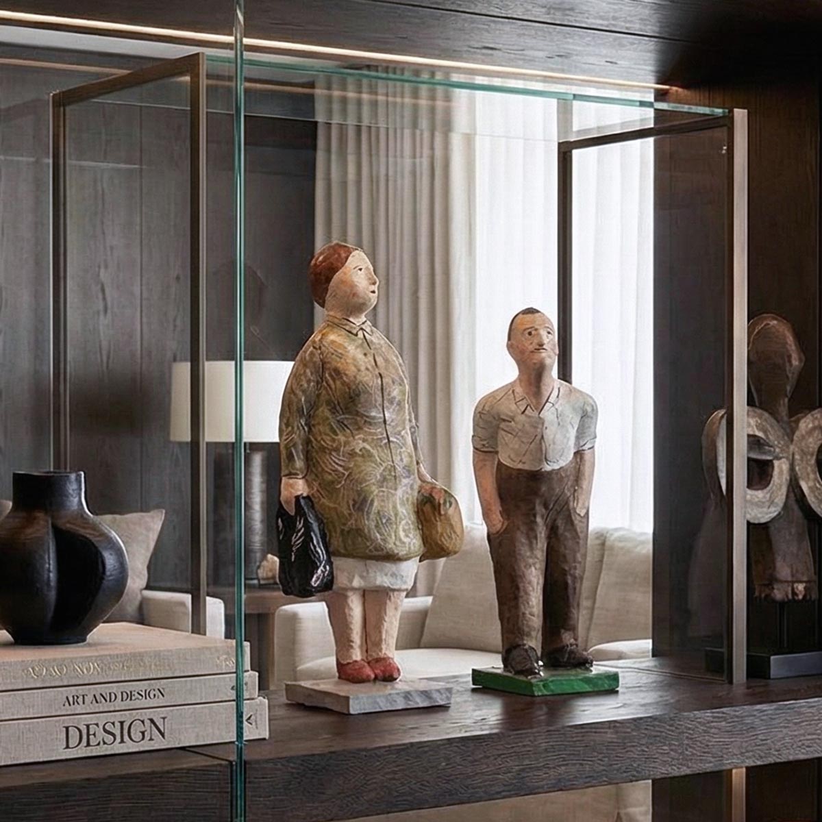 Coppia di sculture figurative in cartapesta dipinta a mano, esposte come pezzi centrali all'interno di una lussuosa teca in vetro e bronzo con illuminazione integrata. La libreria in legno scuro a tutta parete contiene anche libri di design, un vaso scultoreo e manufatti etnici in legno e metallo, in un salotto elegante.