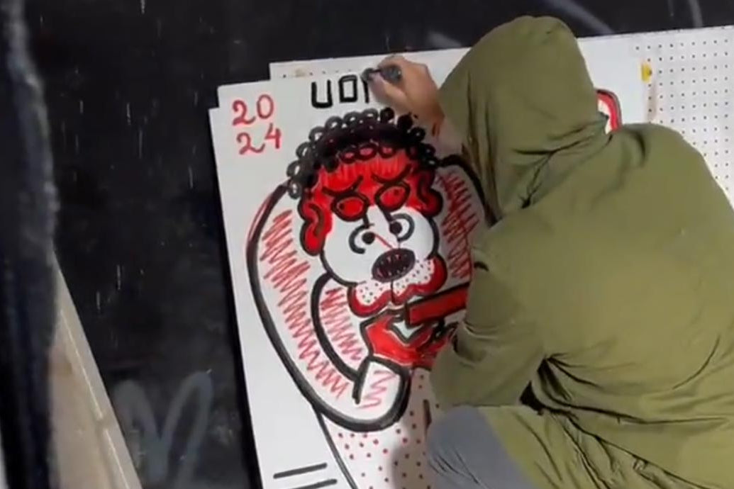 L'artista Uodi, con indosso una giacca verde, è accovacciato mentre dipinge a mano libera una grande pantera rossa stilizzata con contorni neri spessi su una tela bianca con griglie, firmata "UODI" in alto per la collezione vicode
