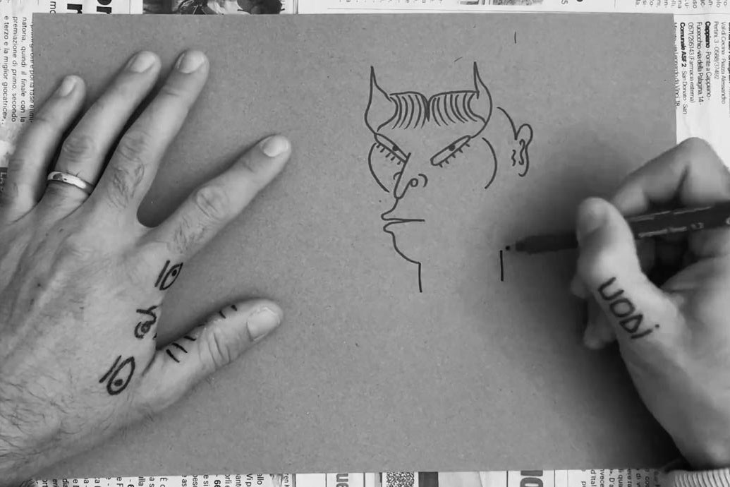 Primo piano delle mani tatuate di Uodi che, con un pennarello nero, tracciano su un foglio di carta grezza il profilo di un personaggio fantastico con corna e grandi occhi, pezzo unico della collezione uodi x vicode