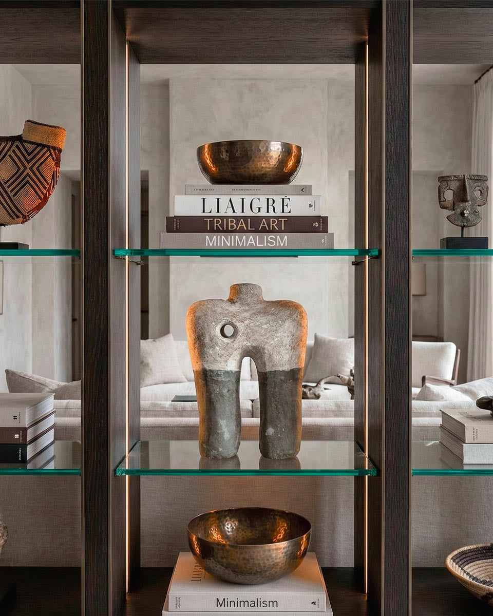 Lussuosa libreria a tutta altezza in legno scuro e bronzo con illuminazione LED integrata in un ambiente living stile Liaigre. Al centro del ripiano in vetro spicca una scultura astratta in cartapesta bicolore (beige e grigio scuro) alta 35 cm con foro centrale, circondata da pile di libri di design ('Liaigre', 'Tribal Art'), ciotole in bronzo martellato e cesti etnici. Sullo sfondo un salotto luminoso.