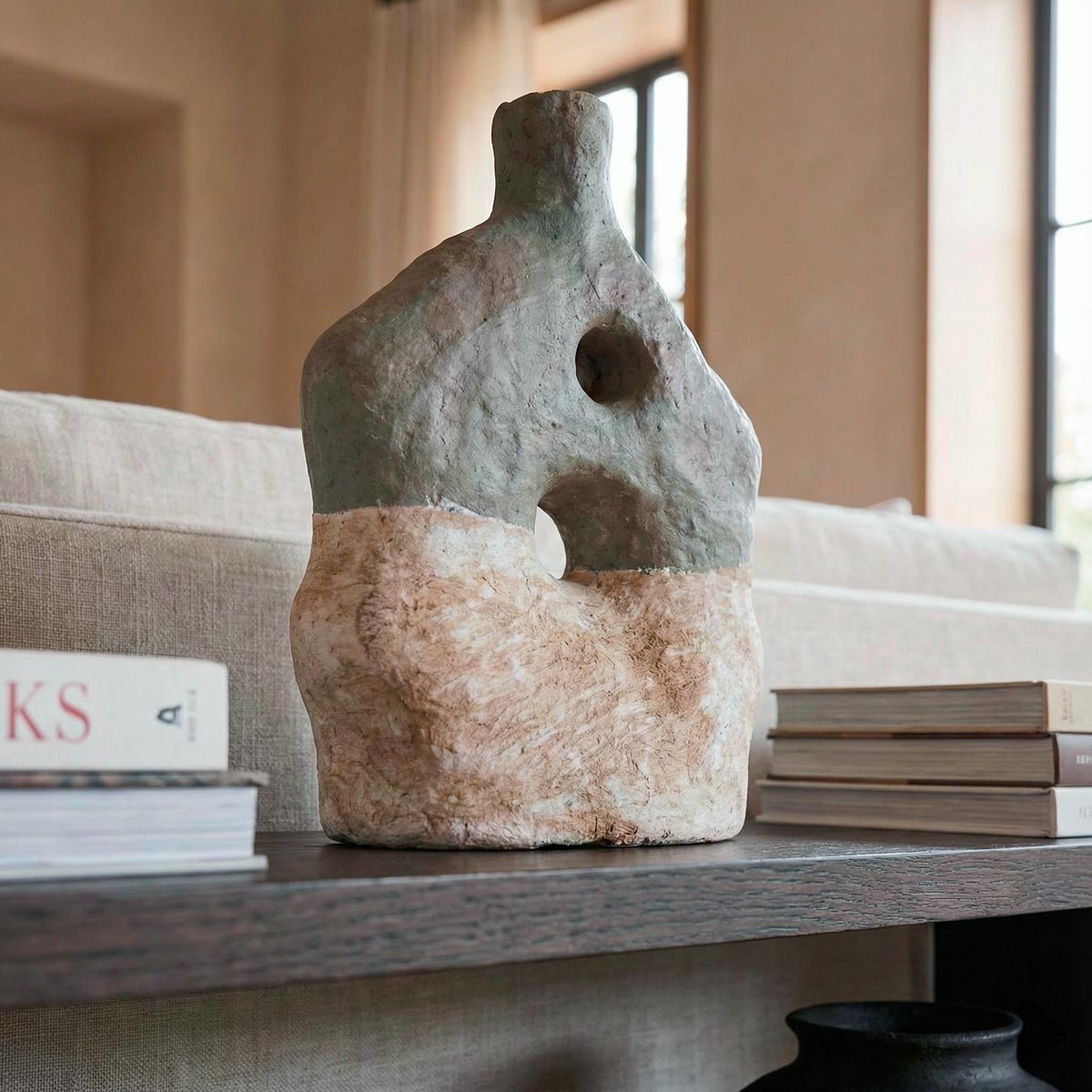 Primo piano con prospettiva leggermente dal basso della scultura astratta in cartapesta. L'inquadratura ravvicinata esalta la texture materica, grezza e artigianale della superficie bicolore e i due fori centrali asimmetrici. La scultura poggia sul bordo della consolle in legno scuro, con i dorsi dei libri e il divano sfocati sullo sfondo per dare profondità.