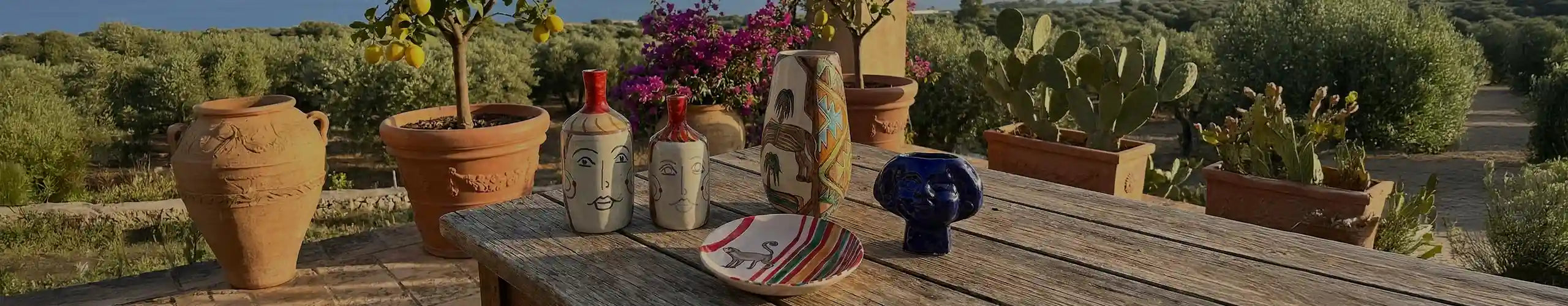 Una collezione di ceramiche siciliane artigianali, inclusi vasi con volti e un piatto con una scimmia, disposti su un tavolo di legno su una terrazza con vista mare, ulivi e piante di agrumi.