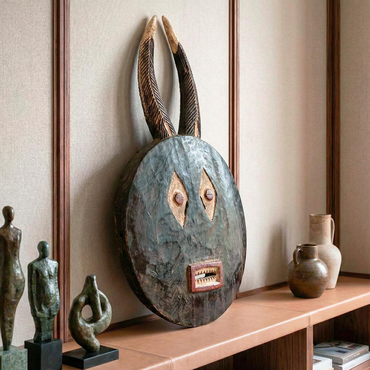 Primo piano dettagliato della maschera tribale in legno tropicale intagliato a mano, che evidenzia la texture materica e le corna. L'oggetto poggia su una mensola rivestita in cuoio naturale, con sfondo sfocato di sculture e parete in tessuto, esaltando l'artigianato di pregio.