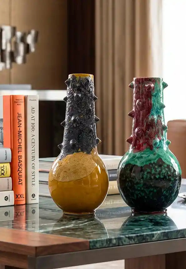 Vasi di design Vicode in ceramica con silhouette a spine e finitura lucida effetto colato nei colori ocra e blu e rosso e verde, appoggiato su un tavolino in marmo accanto a libri d'arte.