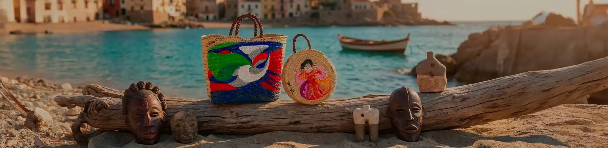 Composizione in stile mediterraneo al tramonto con due borse in paglia Vicode dipinte a mano (una shopper con motivo uccello, una tonda con figura), appoggiate su un tronco in spiaggia e circondate da maschere etniche e sculture in ceramica.