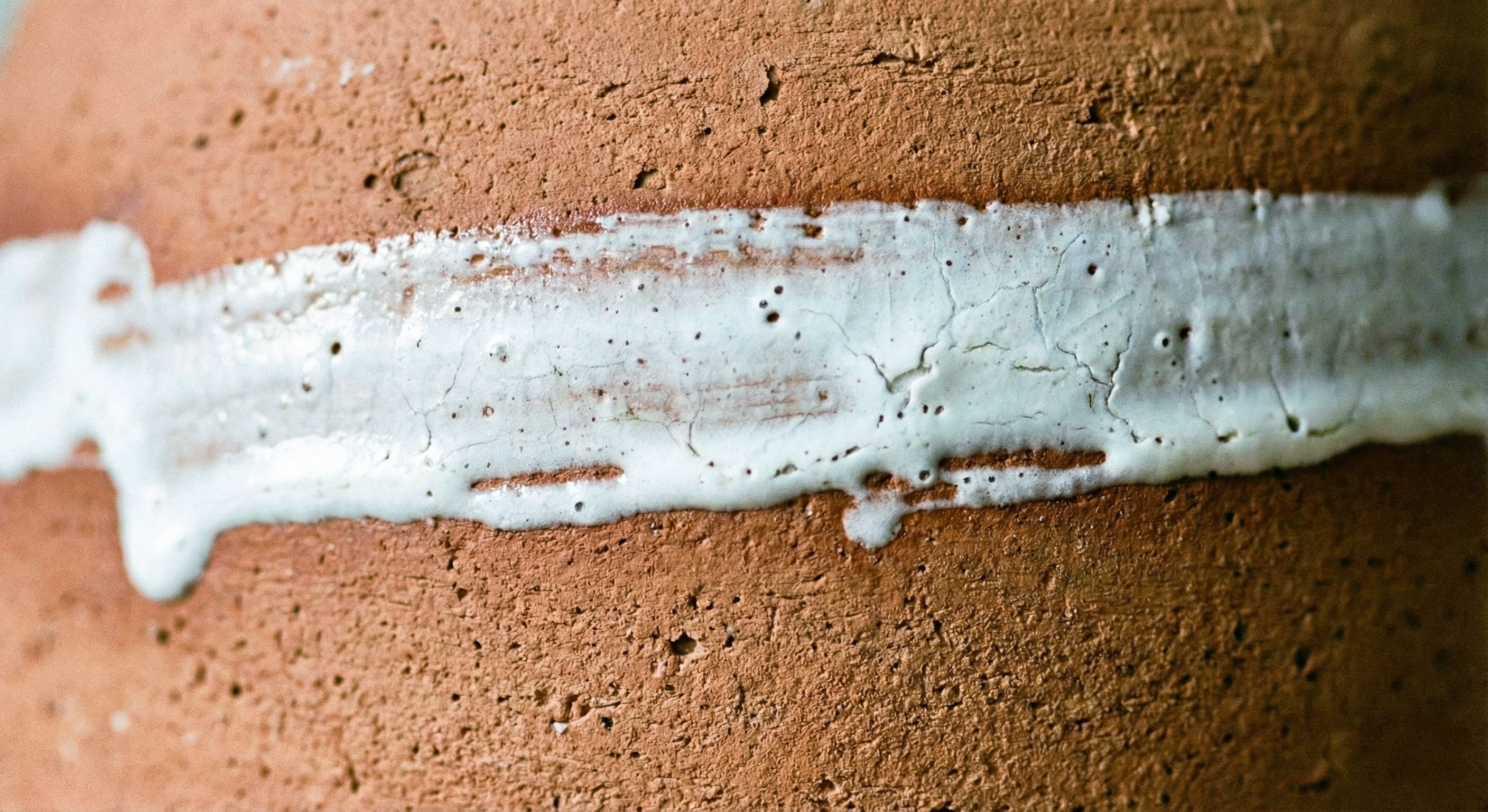 Primo piano macro della texture rugosa della terracotta naturale attraversata da una striatura di smalto bianco, che evidenzia l'anima della materia e la qualità grezza della ceramica.