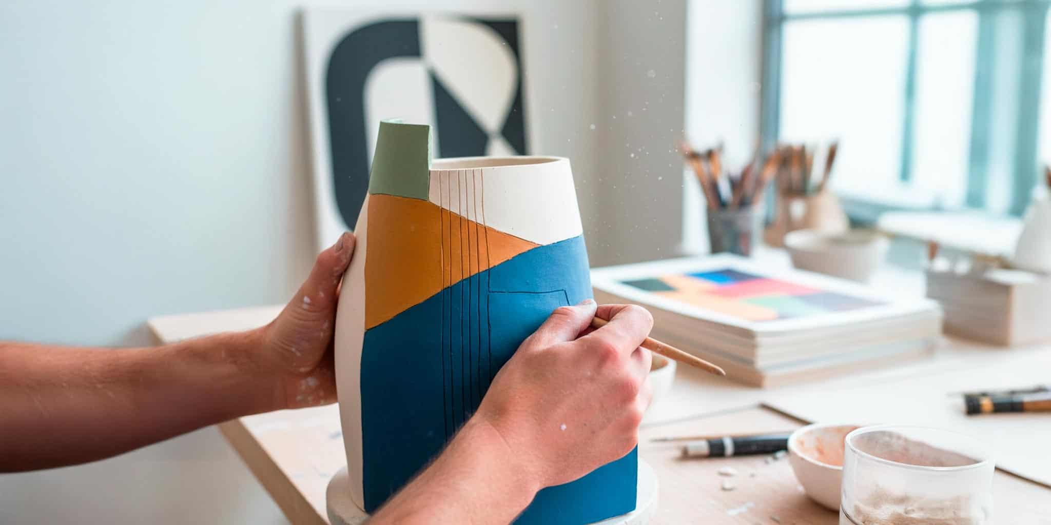 Mani di un artigiano che decorano un vaso in ceramica dal design geometrico moderno con colori blu, verde e terracotta, in uno studio d'arte luminoso.