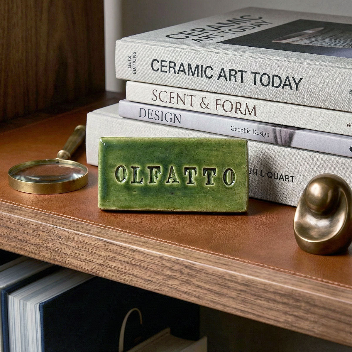 Scultura in ceramica smaltata verde con incisa la parola "OLFATTO" posizionata su una libreria di design accanto a libri d'arte, esempio di decorazione d'interni moderna e sofisticata.