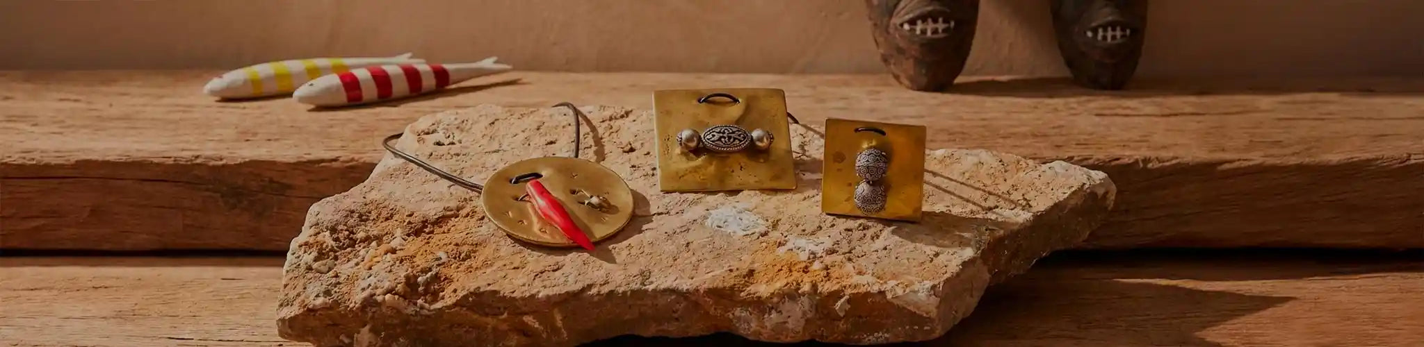 Composizione artistica in stile Architectural Digest che presenta tre monili scultura artigianali in ottone grezzo (un pendente tondo con corno rosso, due pendenti quadrati con sfere in argento lavorato), appoggiati su una lastra di pietra ruvida. Sullo sfondo sfocato, maschere etniche in legno e pesci in ceramica su un tavolo di legno antico.
