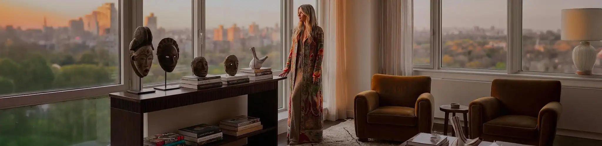Una donna elegante indossa un lungo kimono artigianale a motivi tapestry della collezione Nomad Chic di Vicode. È in piedi in un lussuoso appartamento con vista panoramica sul parco al tramonto, circondata da maschere africane, vasi in ceramica e arredi di design globale.