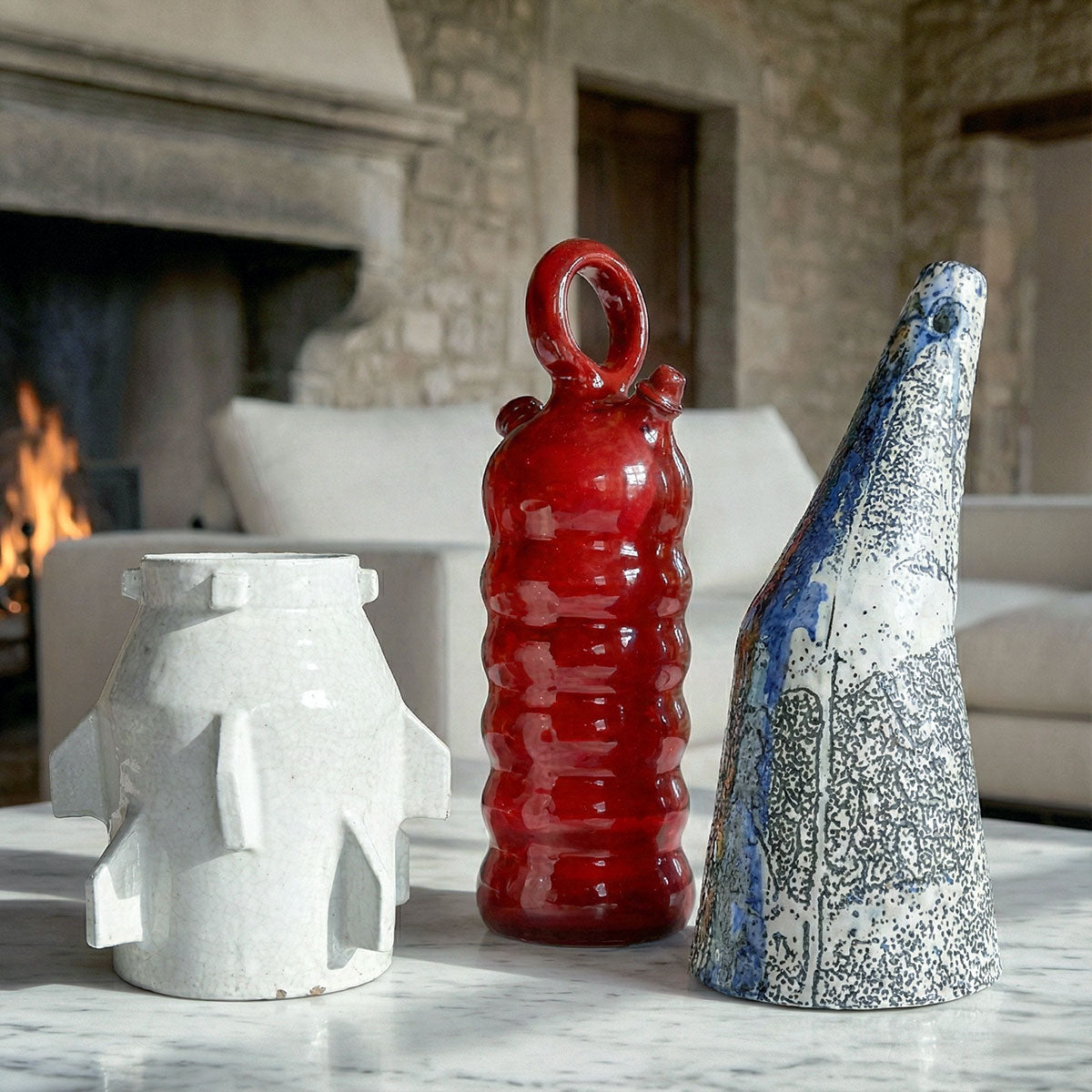 Trio di vasi artistici unici su tavolo in marmo: un vaso bianco geometrico, uno rosso lucido organico e uno blu asimmetrico, in un ambiente rustico elegante.