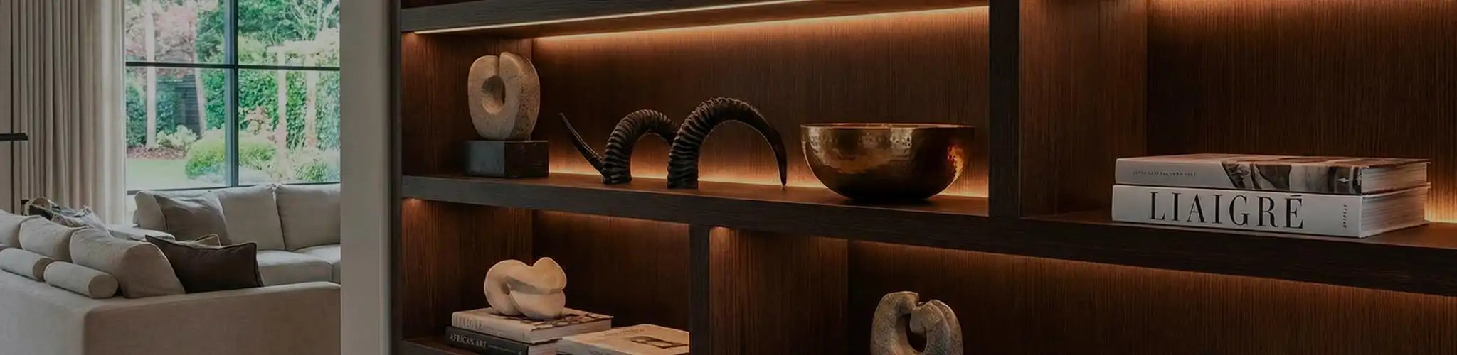 Sofisticata ambientazione d'interni che mostra una selezione curata di decorazioni artigianali su una console in legno scuro contro una parete beige in calce. Include un grande vaso in ceramica testurizzata, una scultura astratta in bronzo e pile di libri d'arte sotto una luce naturale morbida.