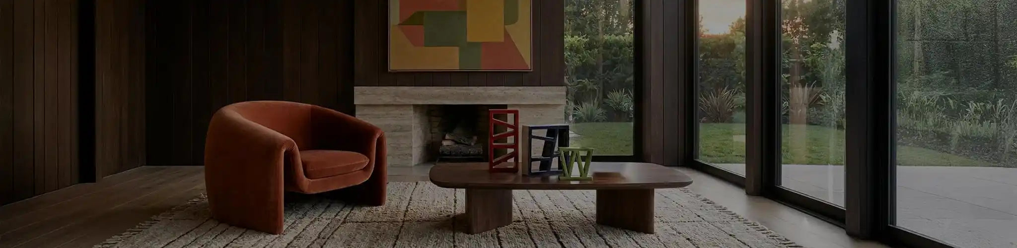 Lussuoso soggiorno in stile anni '70 con rivestimenti in legno scuro e camino in travertino. Una poltrona curva in velluto color ruggine è accostata a un tavolino basso su cui poggiano una grande lampada scultorea in ceramica ocra e tre sculture geometriche colorate (rossa, blu, verde) della collezione Vicode. Arte astratta geometrica alla parete.