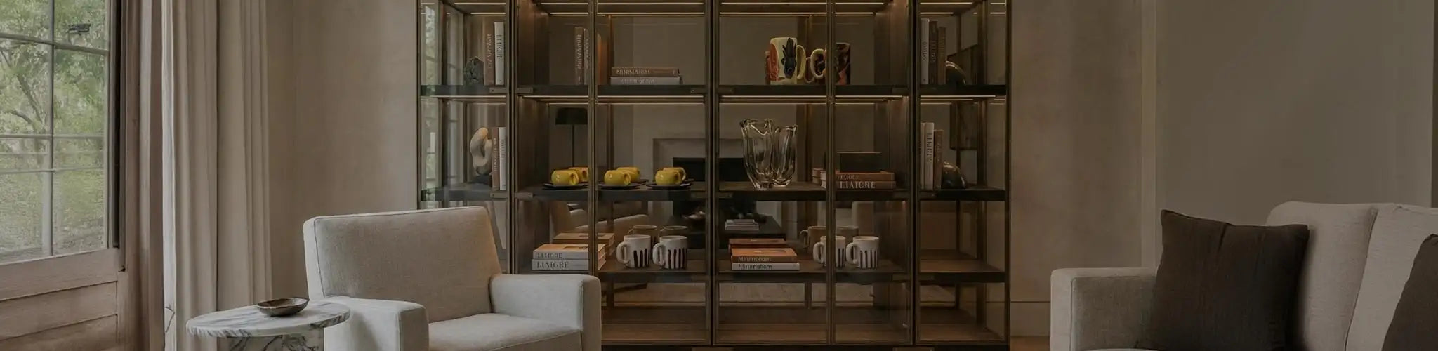 Ampia vetrina a tutta altezza in metallo bronzato e vetro con illuminazione interna a LED, situata in un lussuoso salotto moderno. I ripiani espongono una collezione curata di accessori in ceramica Vicode, inclusi set di tazze gialle, boccali da birra dipinti e mug a strisce, intervallati da libri d'arte e vasi in vetro.