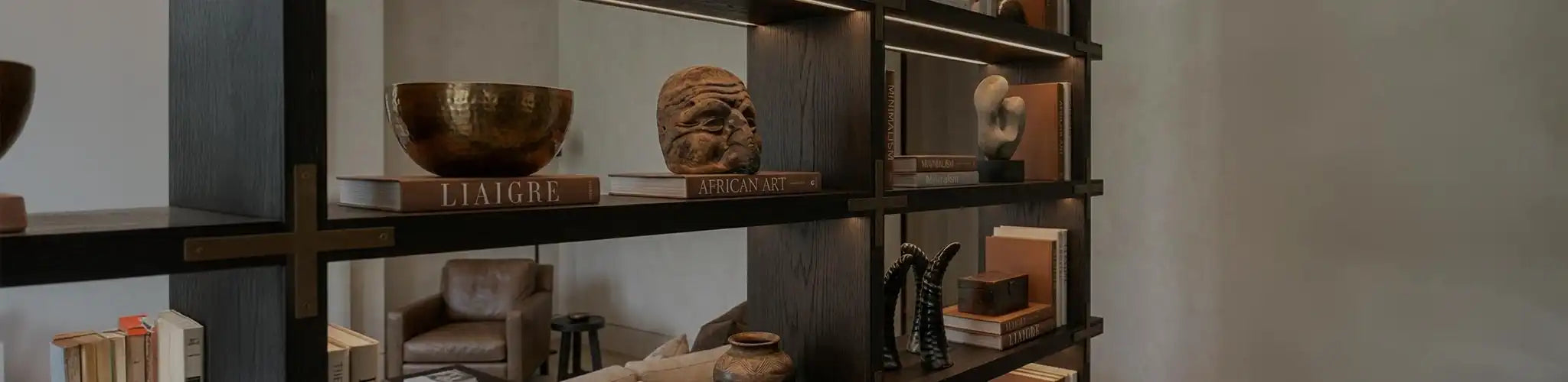 Una sofisticata composizione di oggetti decorativi d'arte su una console in legno scuro. Include una ciotola in bronzo martellato, un'antica maschera di legno su libri d'arte, una scultura astratta in pietra e corni neri decorativi, sotto una luce calda che esalta le texture.