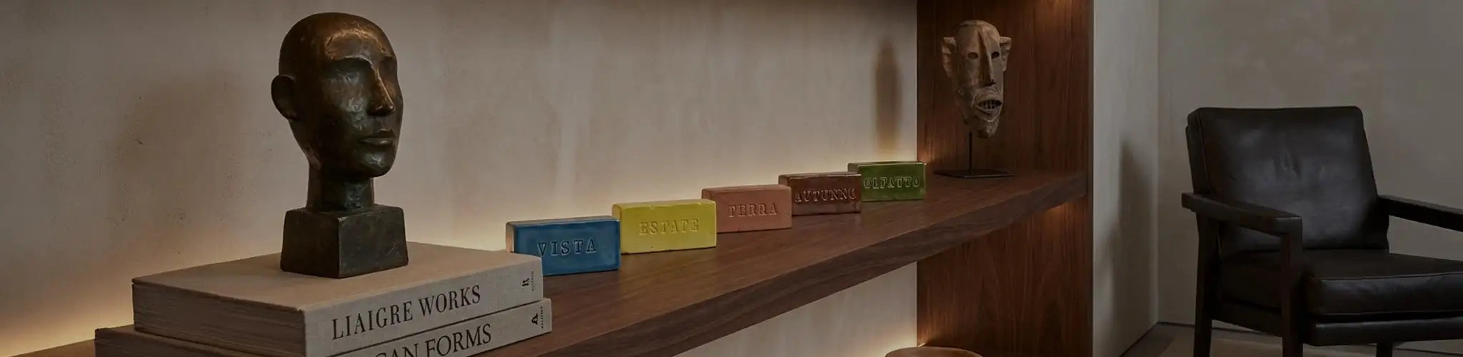 Ambientazione lussuosa con libreria in noce illuminata a LED. In primo piano, una fila di mattoncini in ceramica smaltata 'Edizione Limitata' con incisi i nomi VISTA, ESTATE, TERRA, AUTUNNO, OLFATTO. Sono circondati da una scultura in bronzo, una maschera africana in legno e libri d'arte 'Liaigre Works' e 'African Forms'