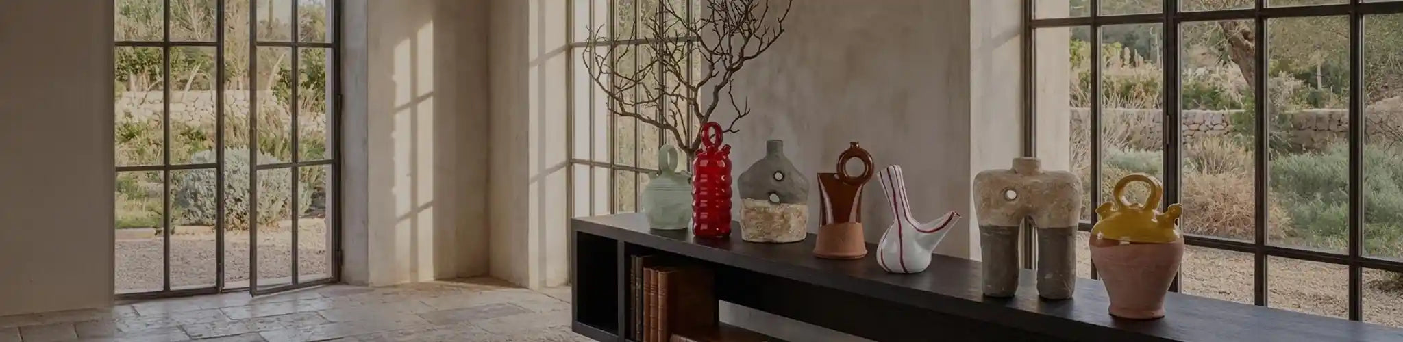 Una collezione curata di botijo in ceramica d'autore Vicode, esposta su una console in legno scuro in un ambiente architettonico in stile Liaigre. La luce naturale proveniente dalle grandi finestre illumina le diverse forme e finiture dei vasi (smaltati, materici, colorati).