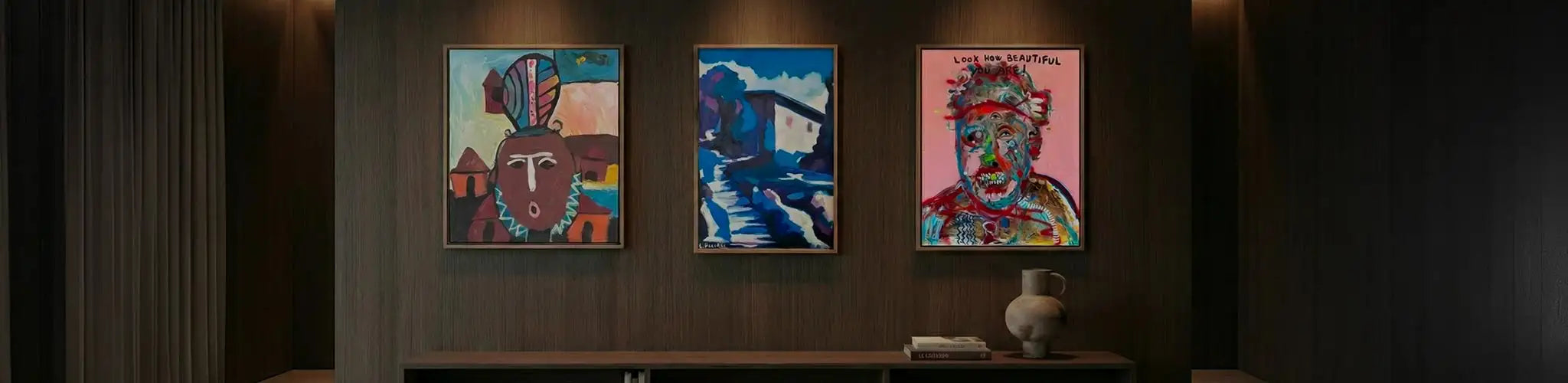 Sofisticata parete galleria in un salotto di lusso Vicode, con rivestimento in legno scuro e illuminazione d'accento che valorizza tre quadri unici incorniciati. Sotto le opere, una console di design con libri d'arte e un vaso in ceramica artigianale.