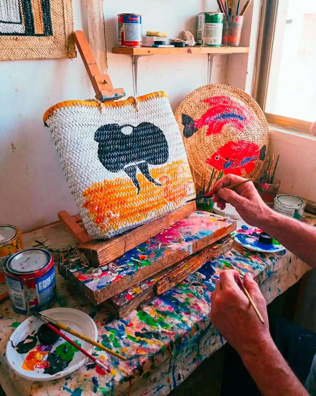 Scorcio luminoso e colorato dell'atelier dell'artista: un banco da lavoro vissuto, colmo di pennelli, colori e attrezzi del mestiere, fa da cornice a una parete dove sono esposte borse di paglia uniche, decorate con vivaci motivi dipinti a mano.