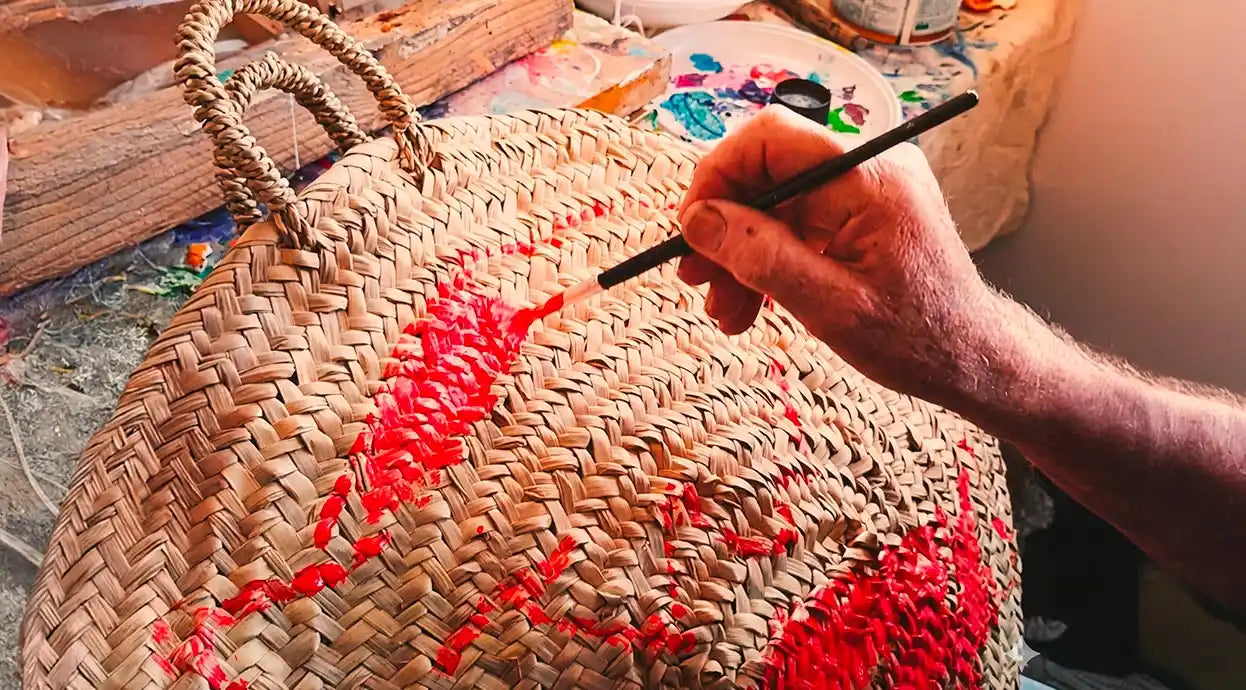 Primo piano delle mani di un artigiano che dipingono con cura un motivo rosso vibrante su una borsa di paglia intrecciata, utilizzando un pennello sottile all'interno del suo laboratorio creativo.