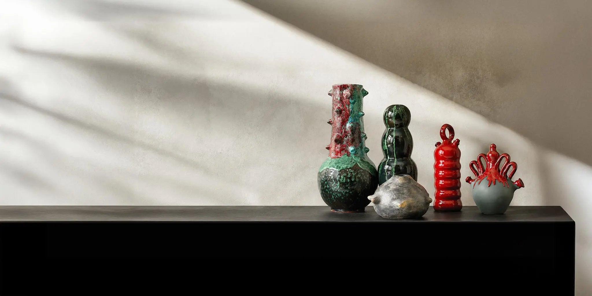 Una selezione curata di vasi artigianali in ceramica e sculture materiche dai toni naturali, disposti su una console in legno scuro. L'illuminazione d'accento calda esalta le texture contro una parete beige lavorata a calce.