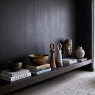 Coppia di sculture artigianali in ceramica, reinterpretazione moderna del 'botijo'. Quella a sinistra è in terracotta opaca, quella a destra in ceramica smaltata bianca con base grezza. Sono disposte su una lunga mensola sospesa in legno scuro, accompagnate da una ciotola in bronzo martellato, pile di libri di design 'Liaigre' e una maschera etnica in legno, contro una parete boiserie scura.