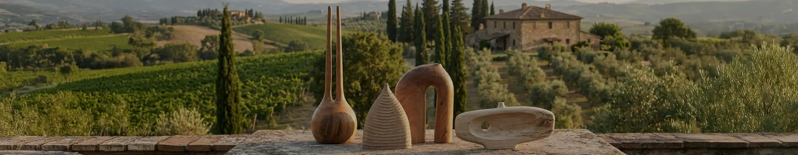 Una fotografia verticale che mostra tre sculture in legno contemporanee – una forma conica rigata, un grande arco liscio e un vaso alto a due colli – disposte su un antico tavolo di pietra. Sullo sfondo, un paesaggio toscano con un casale, cipressi e vigneti illuminati dalla luce calda del tramonto.