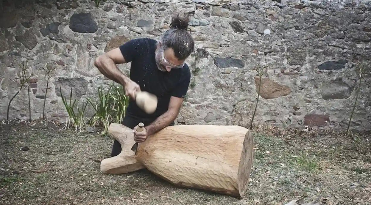 Estebanland seduto nel suo laboratorio all'aperto, circondato da sculture in legno in lavorazione.